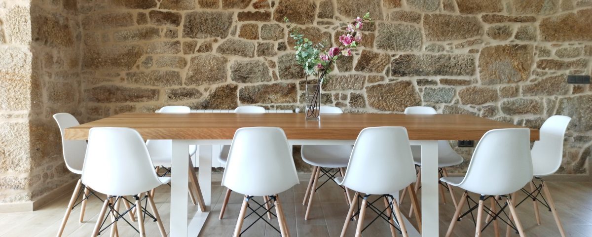 Mesa de comedor en madera con patas blancas y sillas blancas a juego, con un ramo decorativo encima y una pared de piedra de fondo