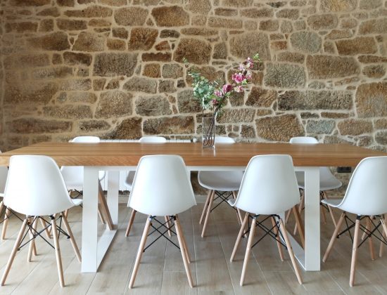 Mesa de comedor en madera con patas blancas y sillas blancas a juego, con un ramo decorativo encima y una pared de piedra de fondo