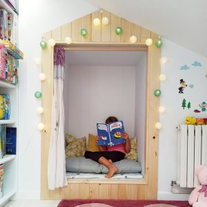 Una niña lee un cuento en su habitación de juegos para niños, decorada con panelado de madera, estanterías y luces