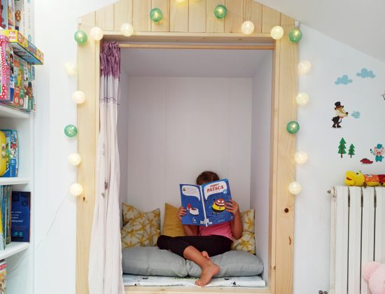 Una niña lee un cuento en su habitación de juegos para niños, decorada con panelado de madera, estanterías y luces